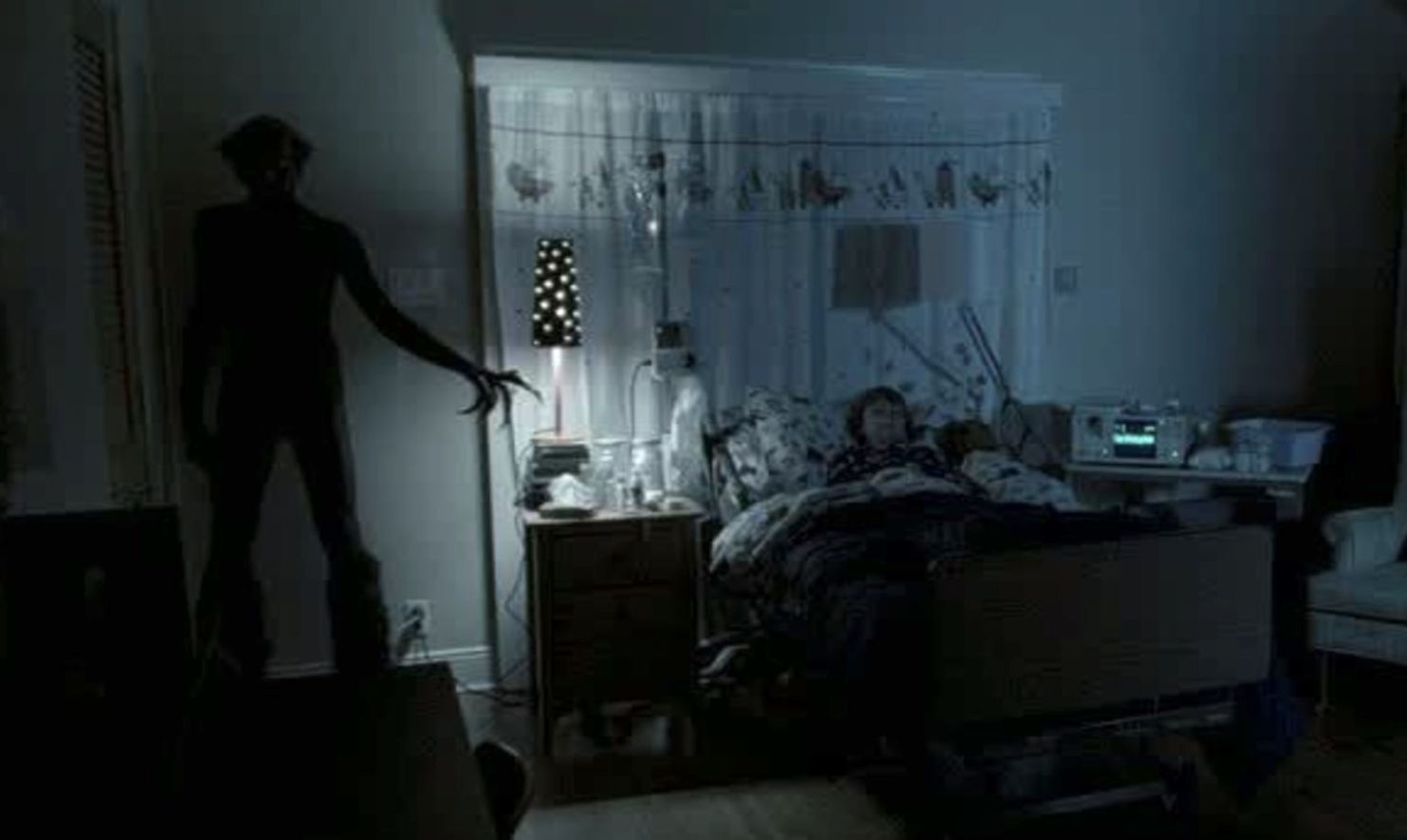 A young boy sleeps in a dark room while a creature stands in the corner