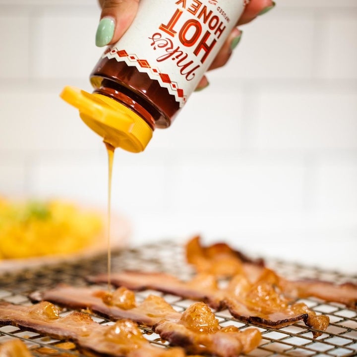 A hand squeezing Mike's Hot Honey onto pieces of bacon on a wire tray