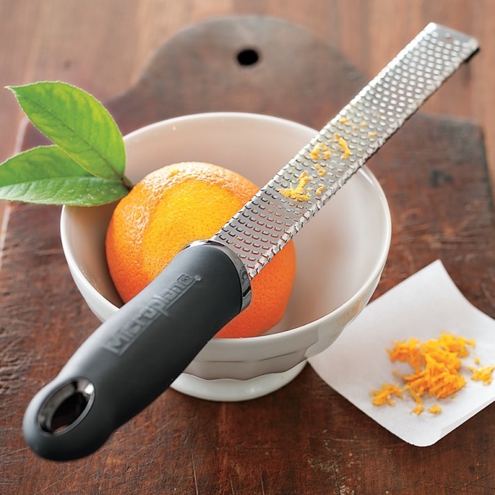 A black and silver grater with orange zest on top, sitting atop a white bowl with an orange