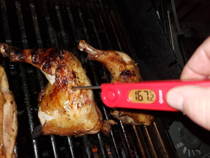 A red meat thermometer reading &quot;167.2 degrees&quot; inserted inside a chicken thigh on the grill