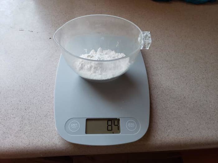 A cup of flour placed on a gray kitchen scale reading &quot;8.4&quot;