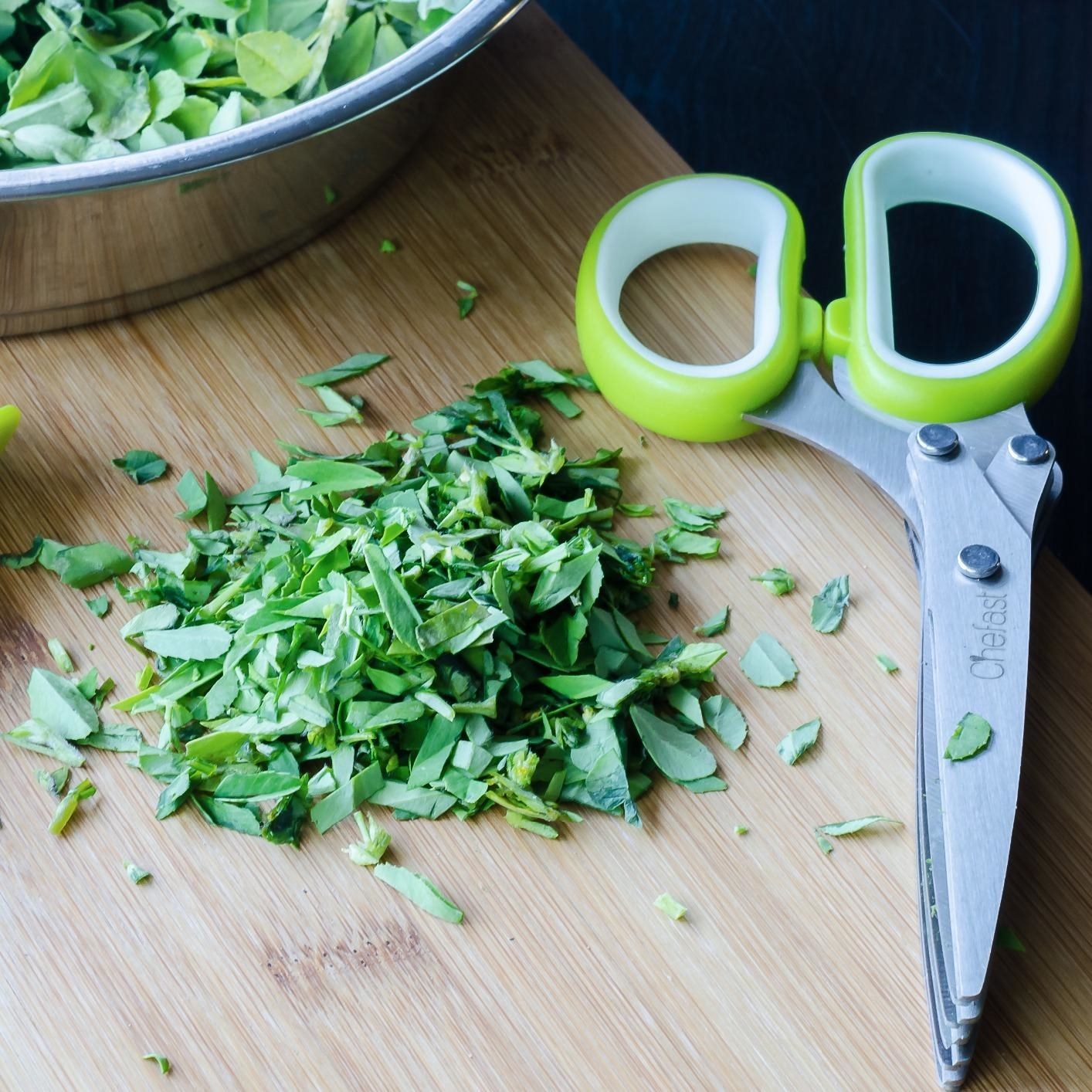 A pair of green herb scissors next to a pile of herbs on a wooden cutting board.