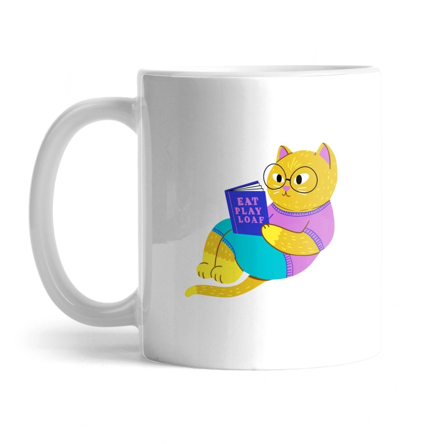 whit mug with a yellow cat wearing glasses and reading a book called &quot;eat play loaf&quot;