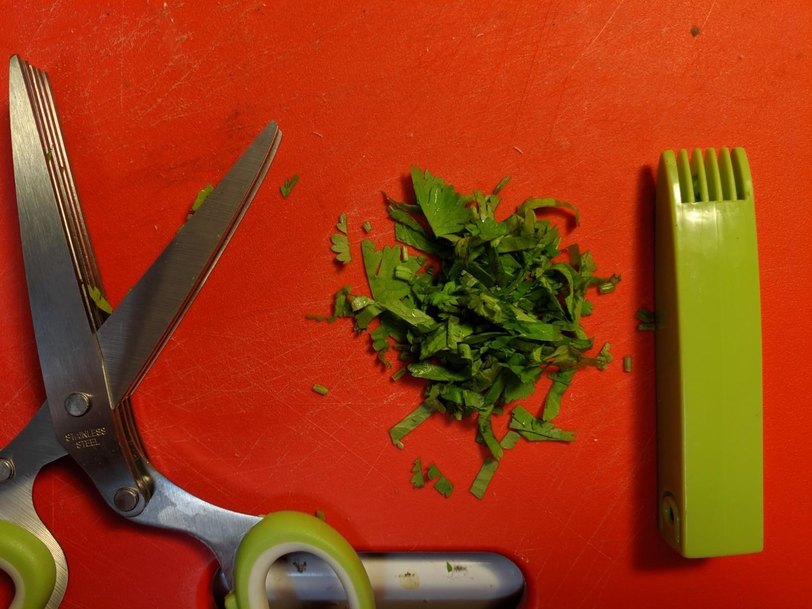 Reviewer&#x27;s scissors and a pile of cilantro cut using them, plus the cover with the cleaning comb built in