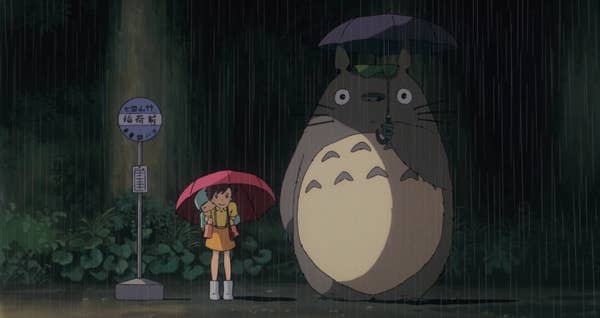 Two girls and a large creature stand in the rain at a bus stop.