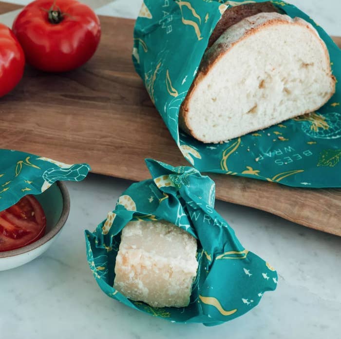 Three teal beeswax storage wraps holding bread, cheese, and a bowl of tomatoes