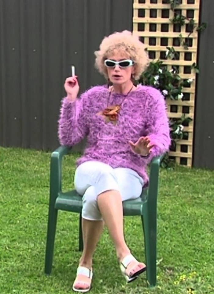 Woman sitting on a plastic lawn year wearing a pink jumper and sunnies, smoking a cigarette