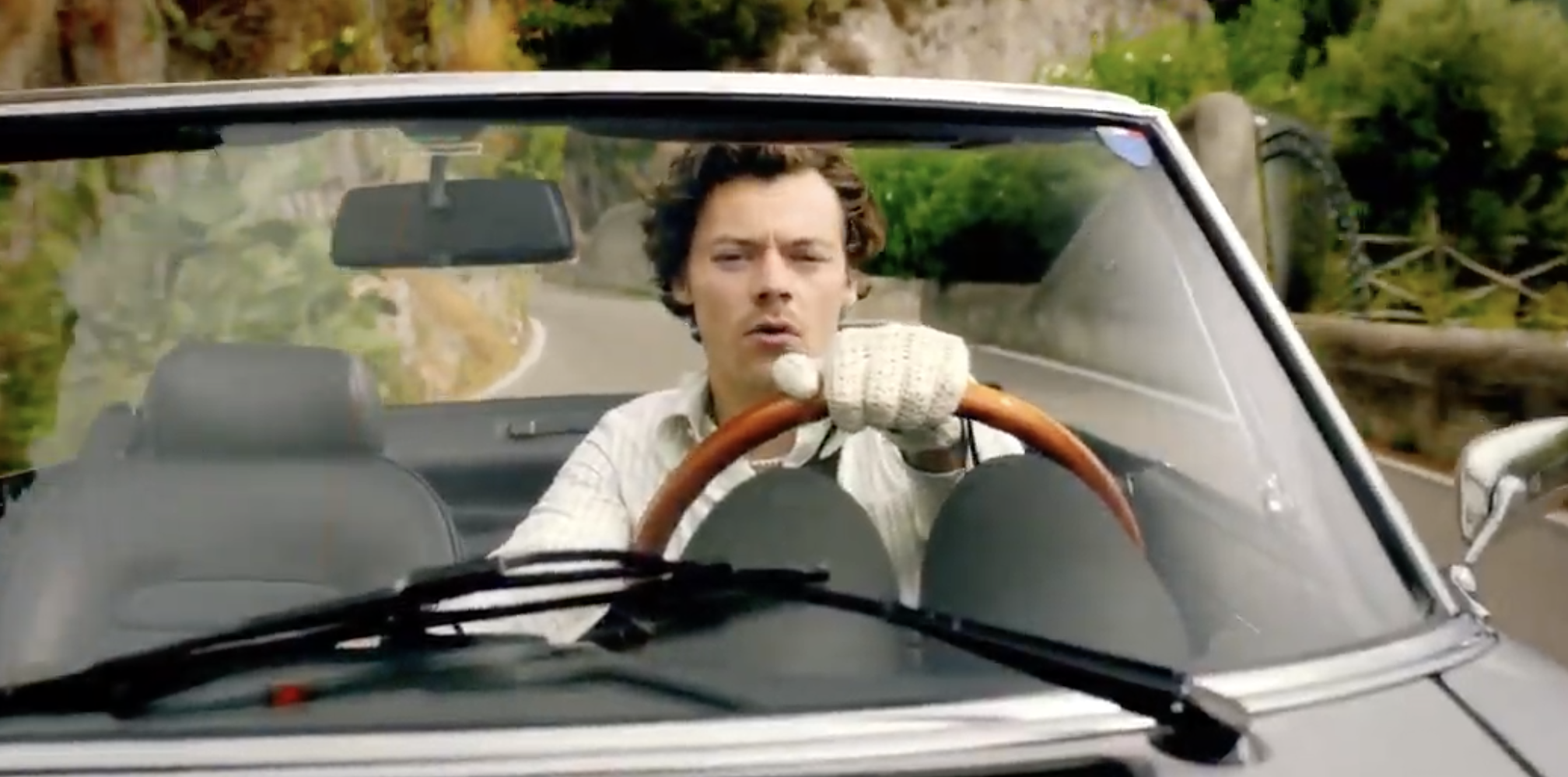 Harry driving a car with the top down while wearing driving gloves
