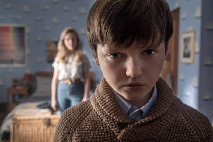 Young child wearing a sweater standing in a bedroom with a woman standing in the background