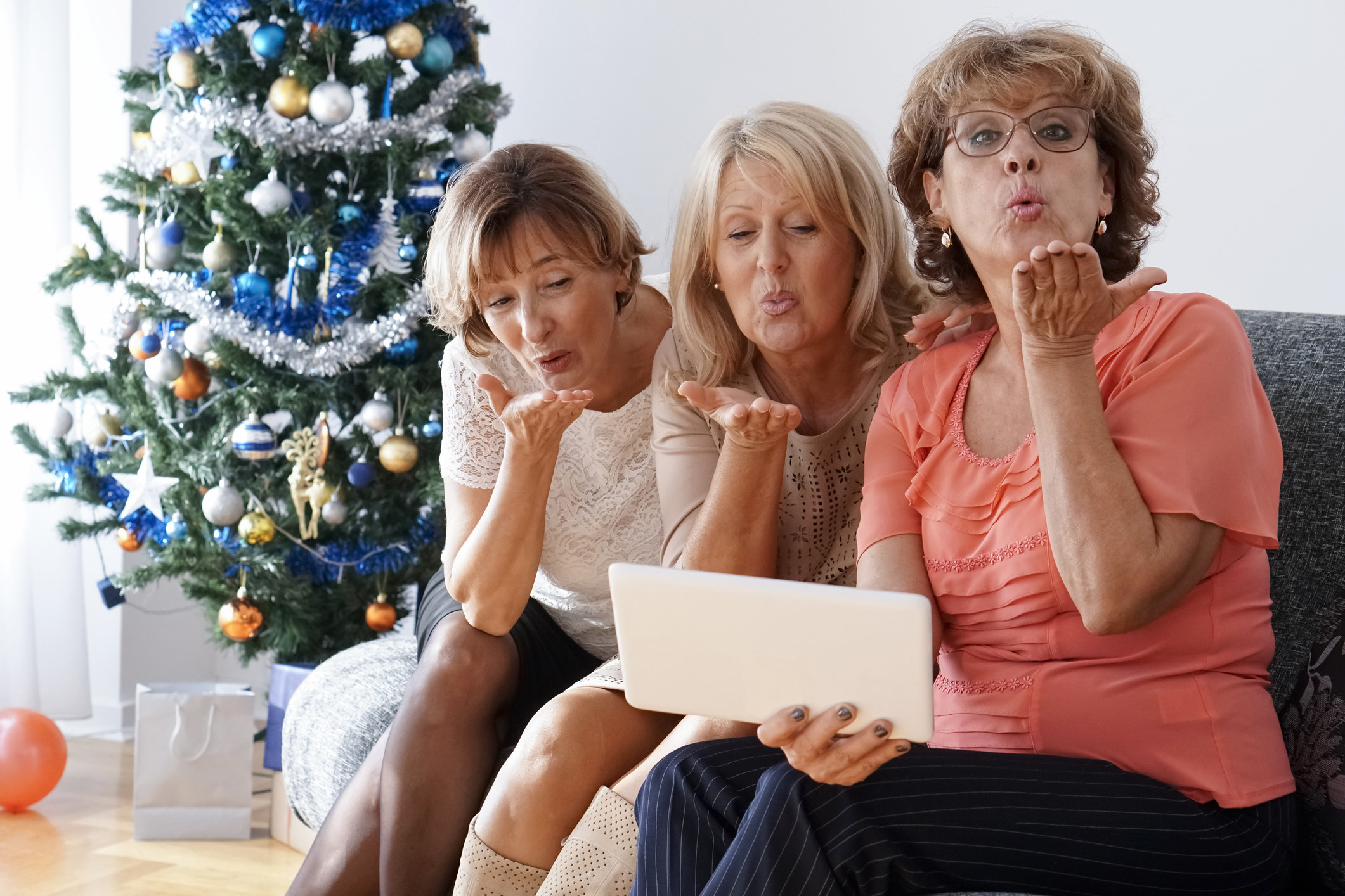 Three women sitting on a couch, with a Christmas tree in the background, blowing kisses