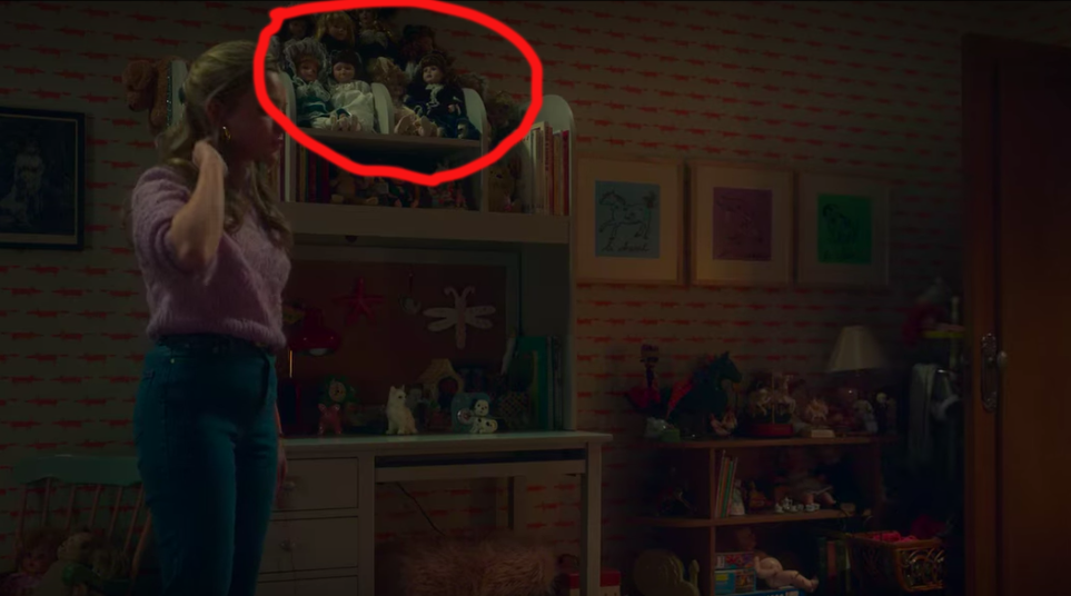 Amelia Bea Smith as Flora standing in a room next to bureau that has a large collection of dolls