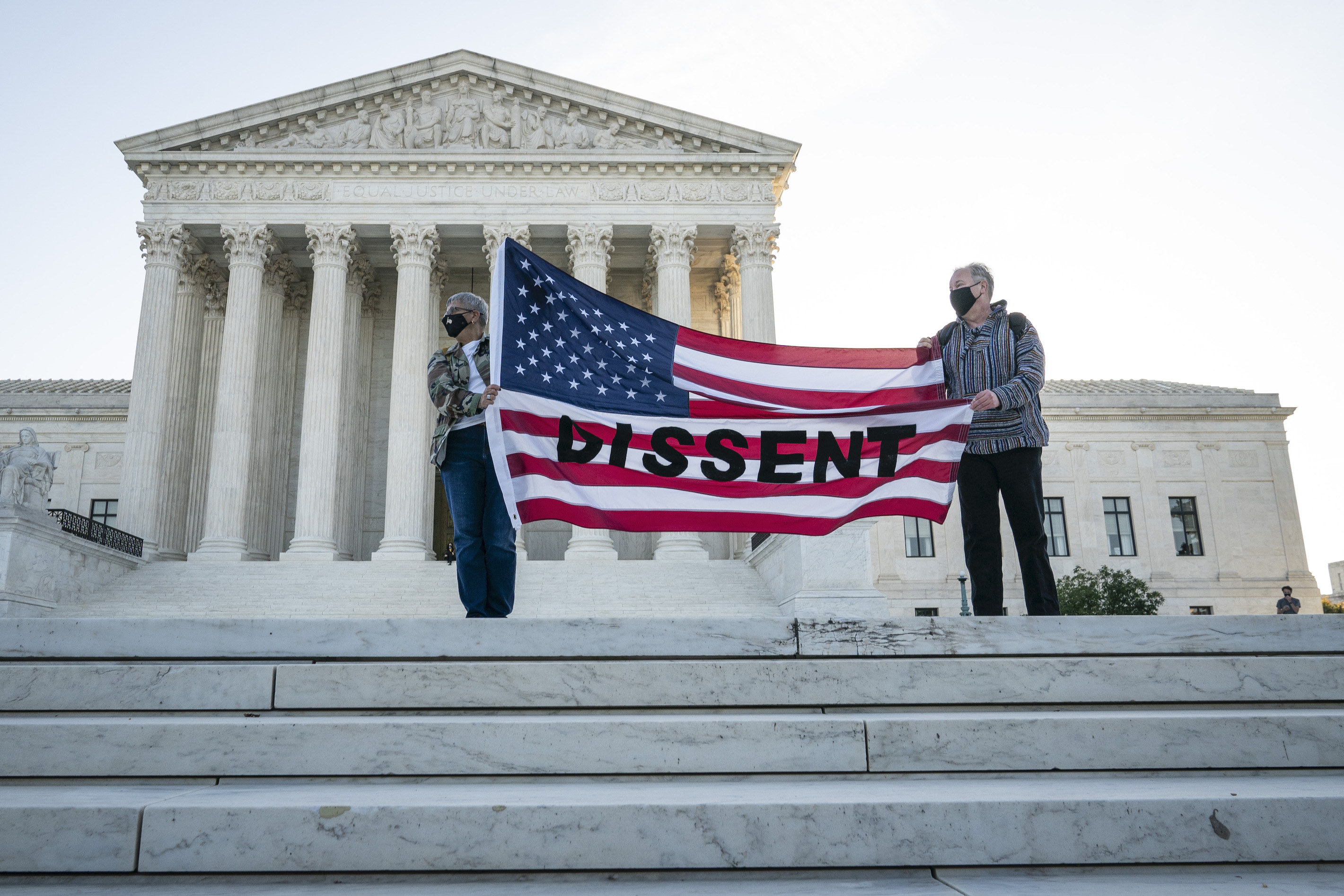 Two people wearing masks holding a large US flag with &#x27;dissent&#x27; written across it as they stand on the steps of the Supreme Court