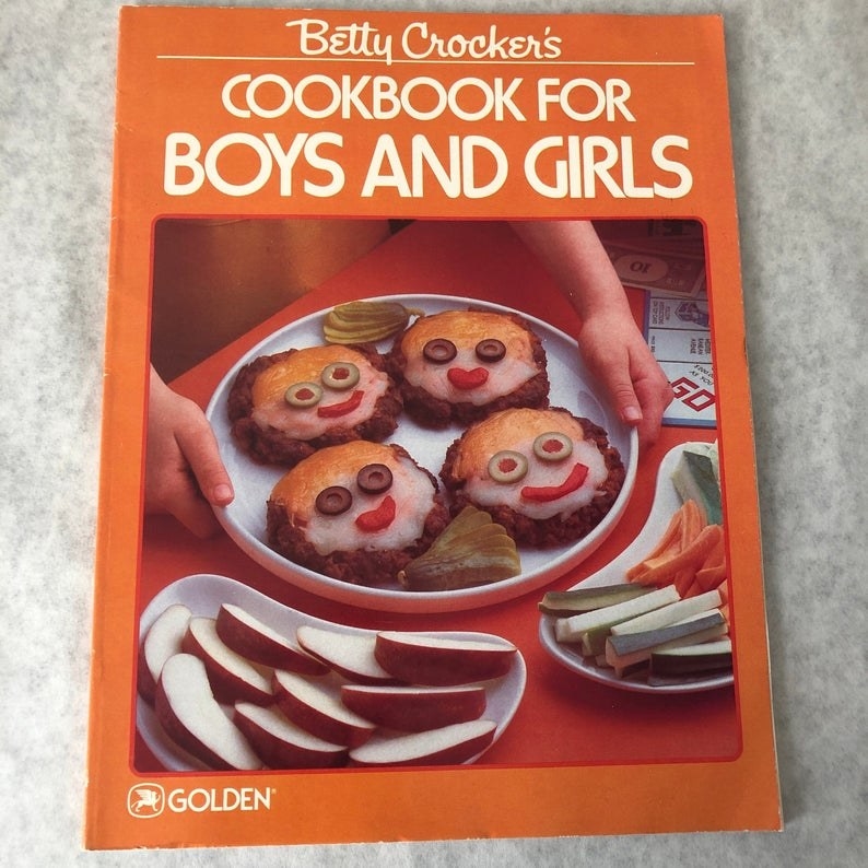The orange cover for Betty Crocker&#x27;s Cookbook for Boys and Girls, which features a hand putting don a plate with for hamburger patties with smiling faces made of cheese and olives