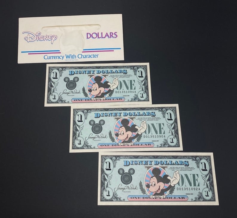 An envelope with Disney Dollars written on it and three $1 Disney Dollars that features Mickey waving on them