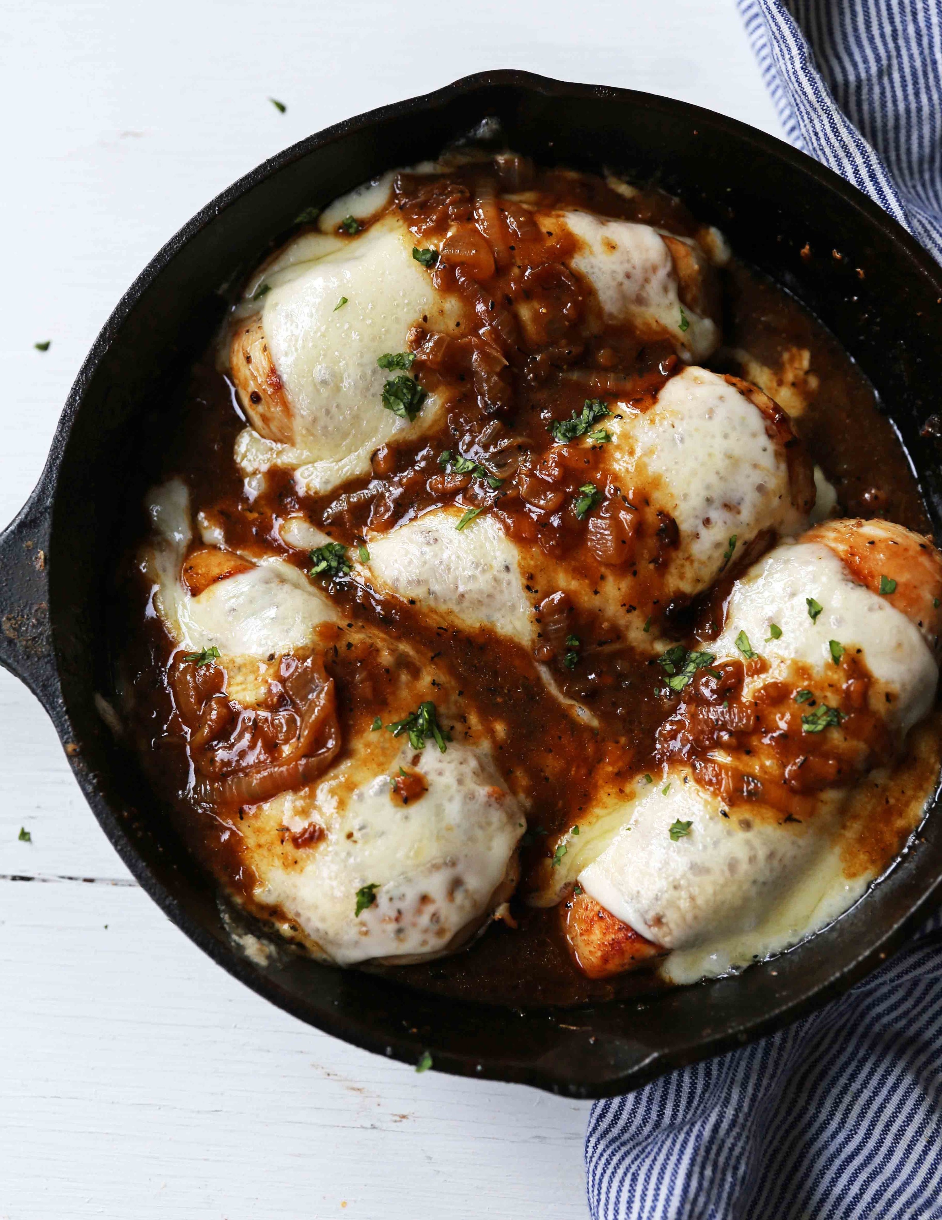 A cast iron skillet with four chicken breasts covered in melted cheese and caramelized onions