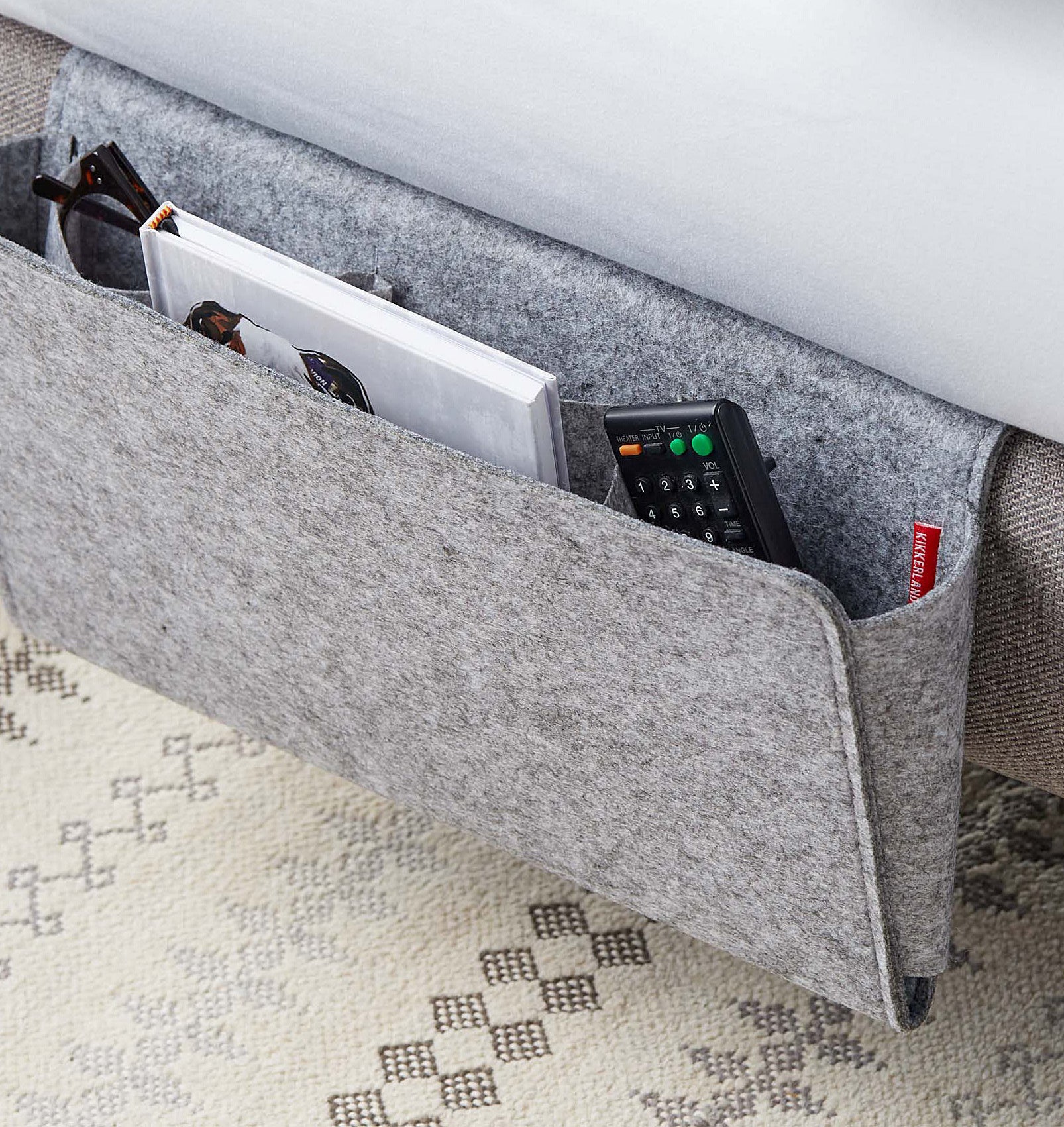 A felt folder hanging from a bed frame with a book, a remote and a pair of glasses organized inside