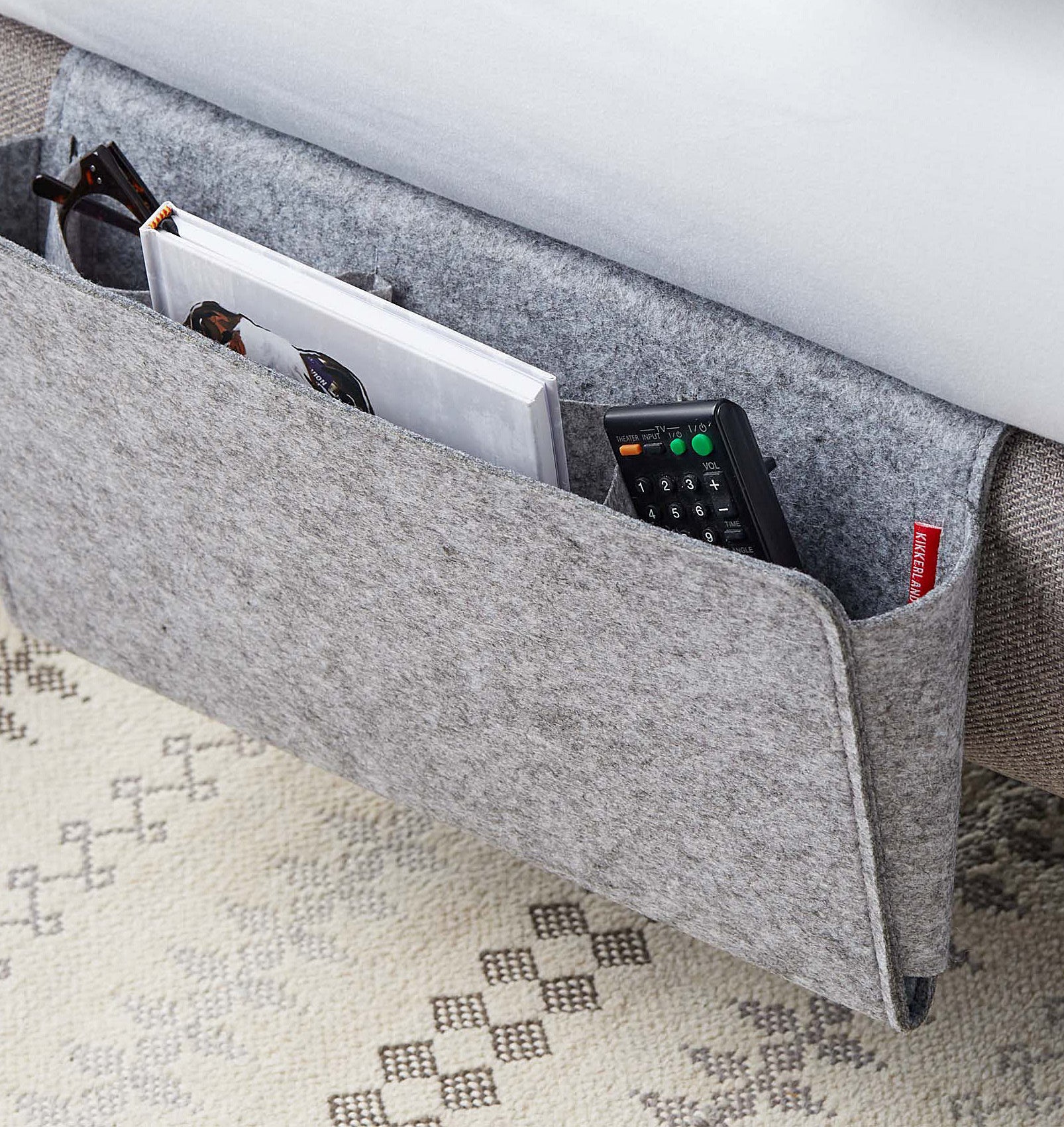 A felt folder hanging from a bed frame with  a book, a remote and a pair of glasses organized inside