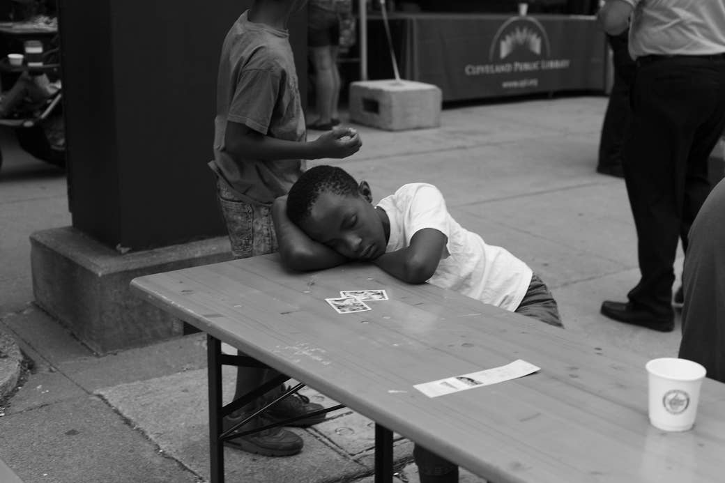 A small boy sleeps at a table outside while other children play in the background