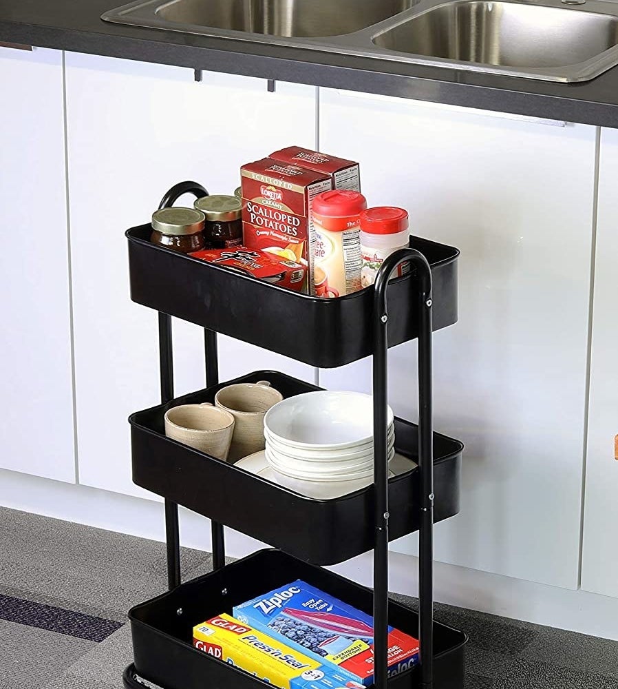 A metal rolling utility cart with spices in one shelf, plates and bowls on the second shelf, and plastic food wrap boxes on the lowest shelf
