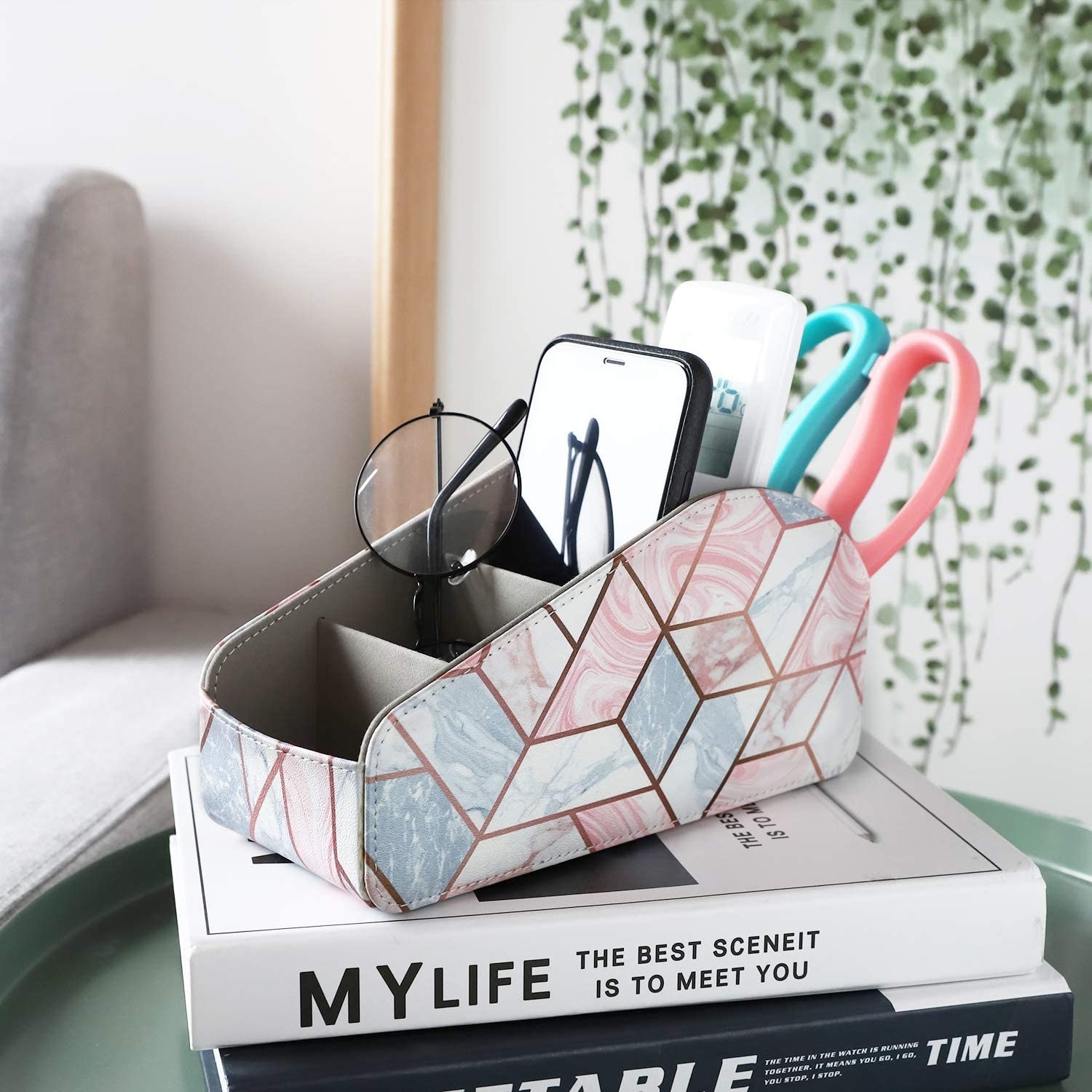 A fabric remote control organizer sitting on a stack of books  There are a pair of glasses, a phone, a remote, and a pair of scissors in a different compartment