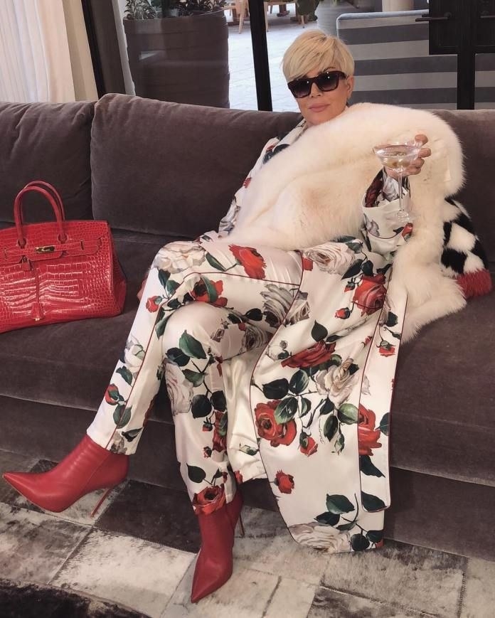 Kris Jenner wearing a luxurious outfit, holding a glass of wine, and posing on a couch