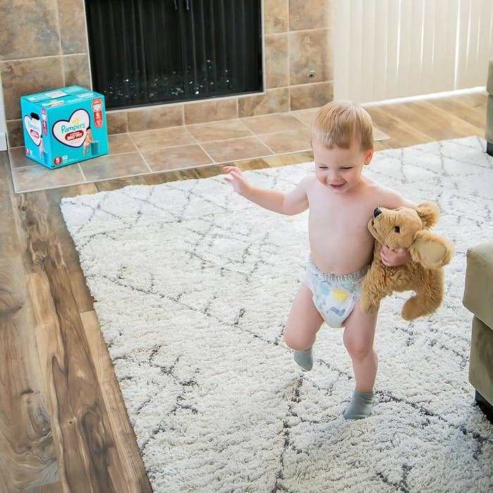 Smiling baby walking across carpet in diaper while holding stuffed dog