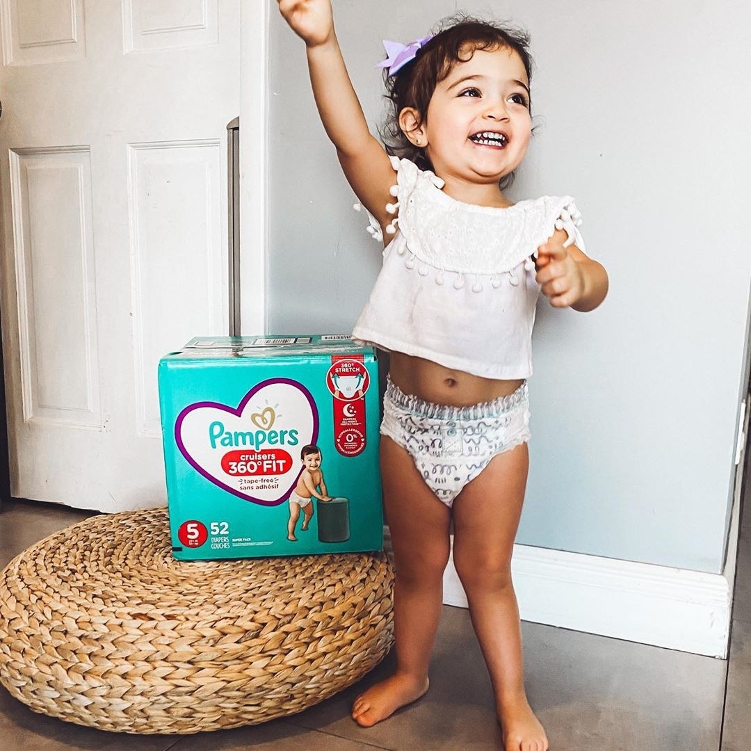 Toddler girl standing and smiling next to box of Pampers® Cruisers 360° Fit™ diapers