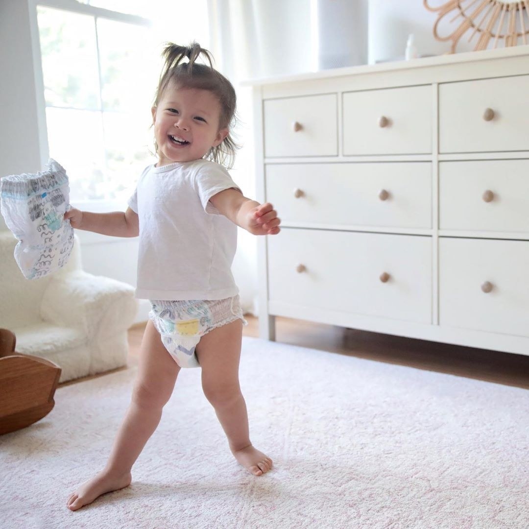 Toddler girl standing in white T-shirt and diaper, holding another diaper, and smiling