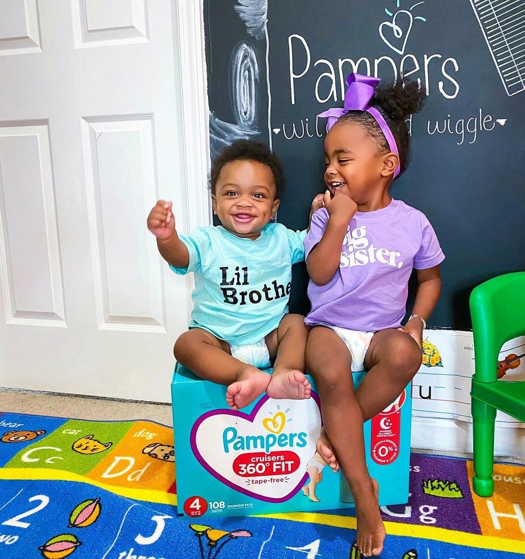 Toddler girl and baby boy sitting on box of Pampers® Cruisers 360° Fit™ diapers and smiling