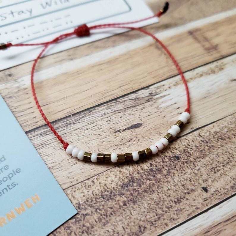 An adjustable red string bracelet with white and gold beads in a morse code letter sequence