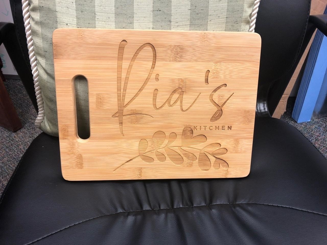 A reviewer showing a wooden cutting board personalized to say &quot;Ria&#x27;s kitchen&quot; with a leaf