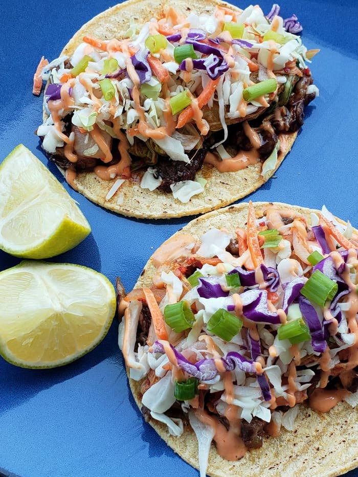 a close up of two Korean tacos on a plate, with bright green scallions, red cabbage, and a Sriracha mayo sauce on top