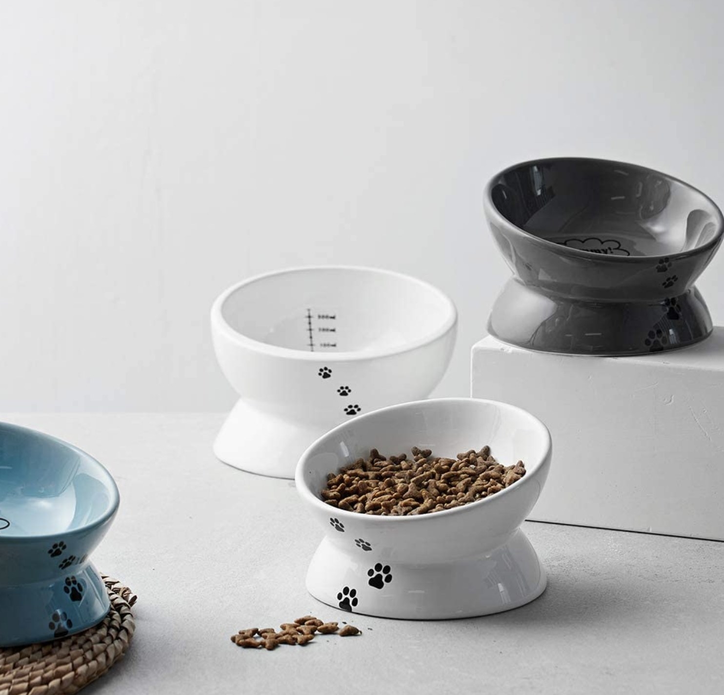 titled cat food bowls with paw prints on the outside of them and measurements on the inside of them. There are two white bowls, one blue bowl, and one grey bowl. One of the white bowls contains dry food