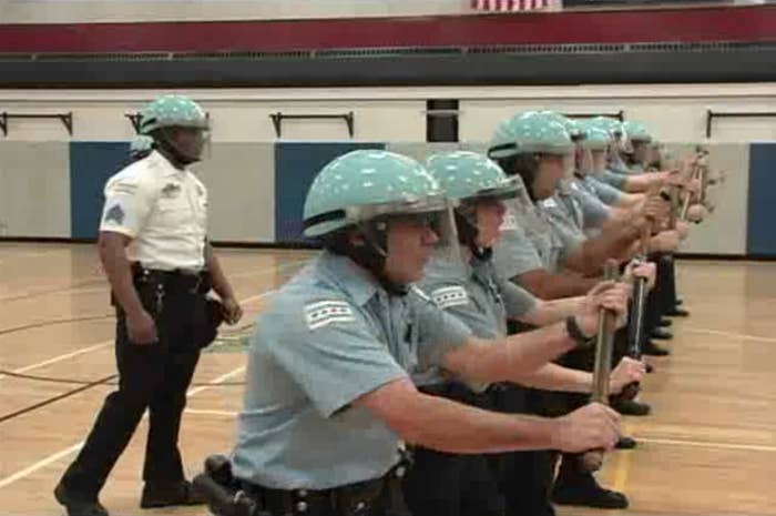 Cops wearing protective gear and holding batons stand in a line in a gymnasium