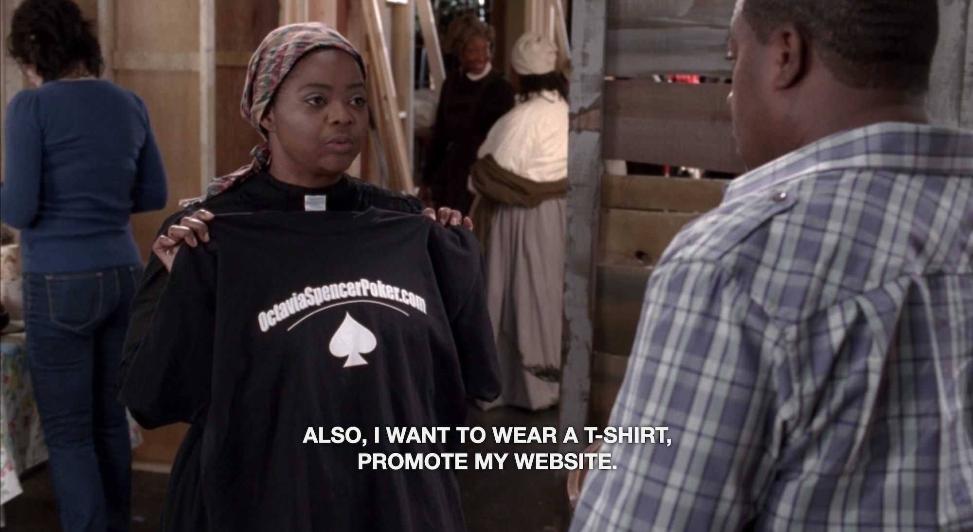A woman in old timey clothes holding up a T-shirt, with the caption text "Also, I want to wear a t-shirt, promote my website"