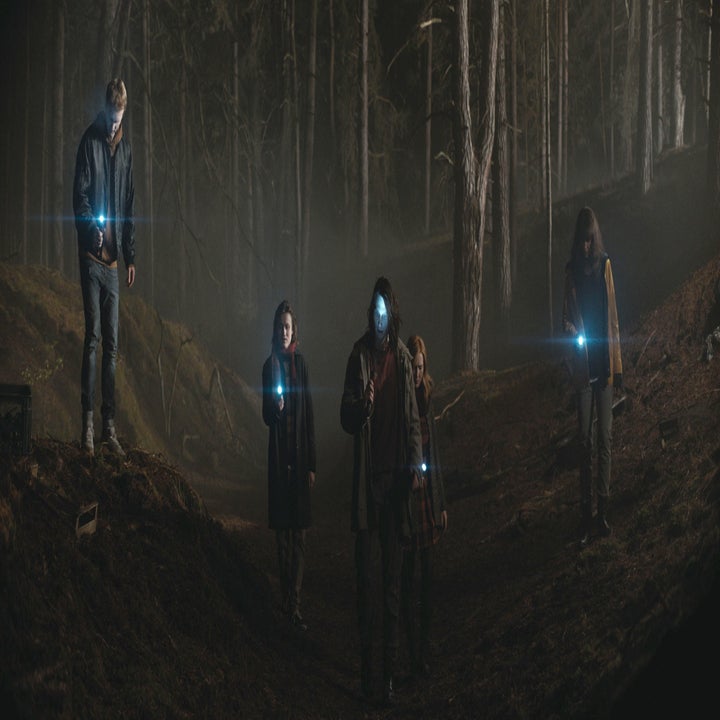 A group stands in a dark, misty forest, their flashlights glowing towards the camera
