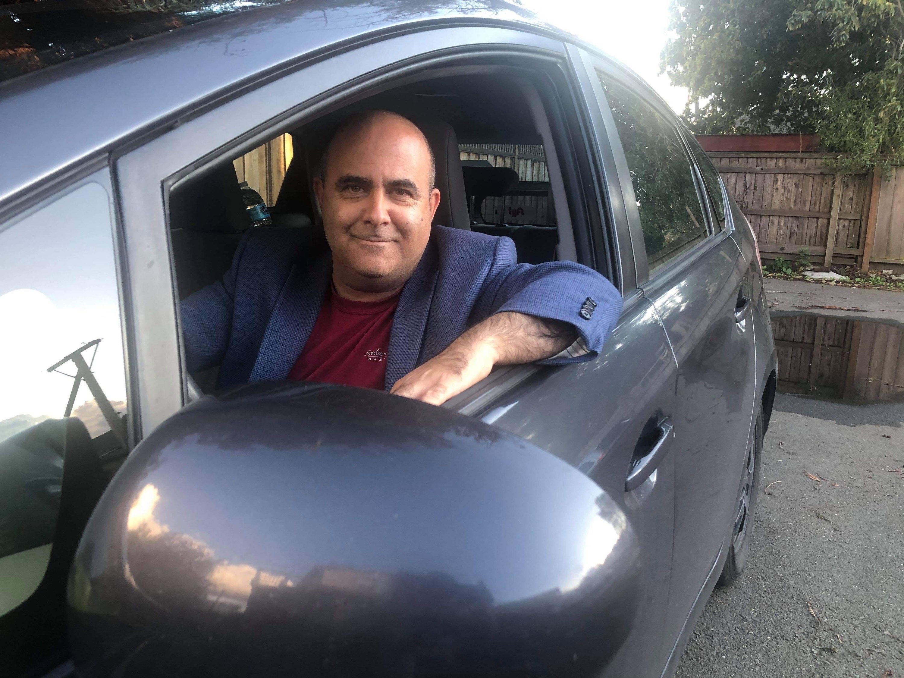 An Uber driver named Edan Alva sits in his car and smiles at the camera