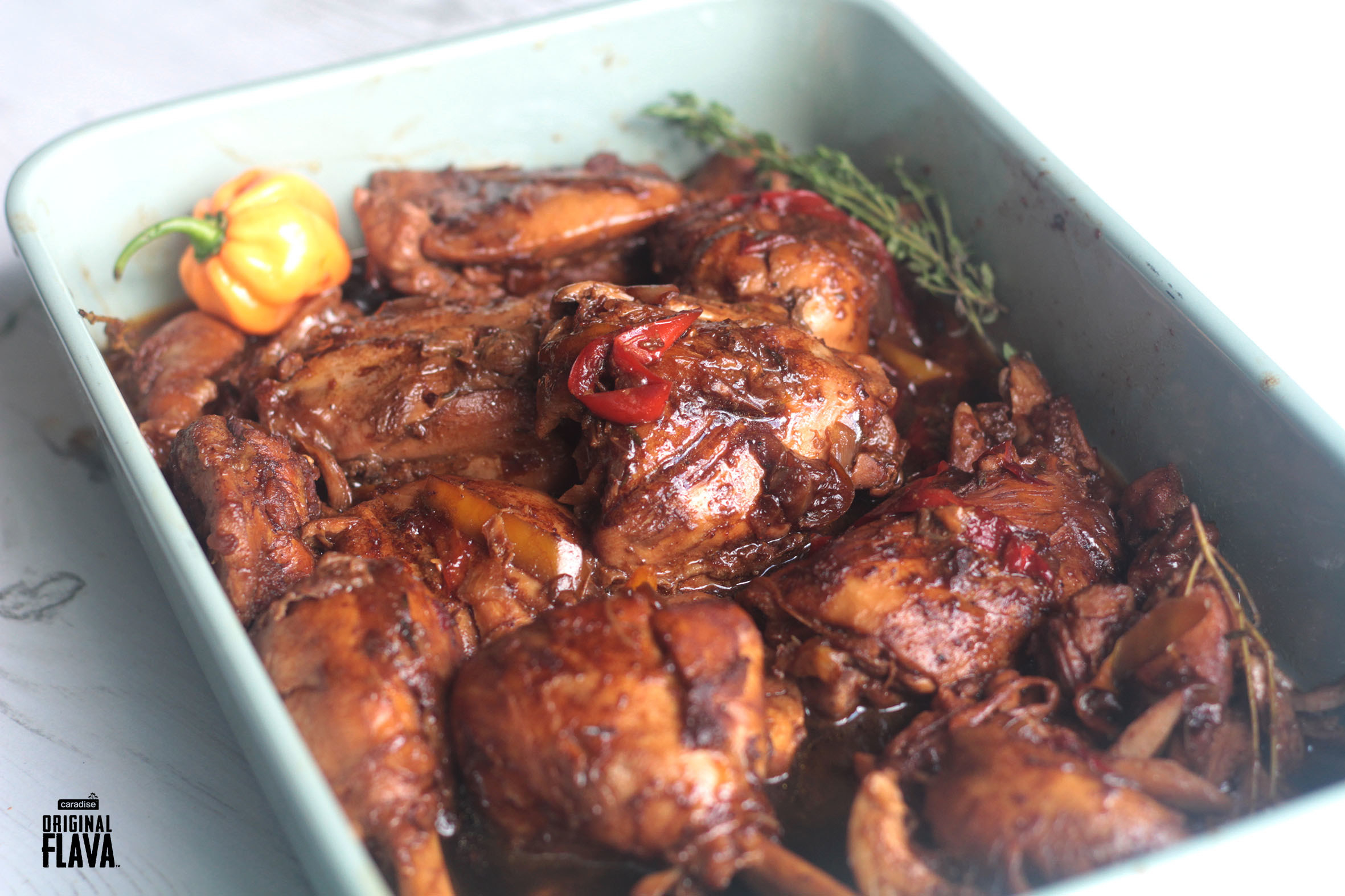 A baking dish of brown stewed chicken thighs and legs garnished with a small scotch bonnet pepper and sprigs of thyme.