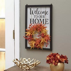 A sign that says "Welcome to our Home" with a hanging autumn red, yellow, and orange wreath