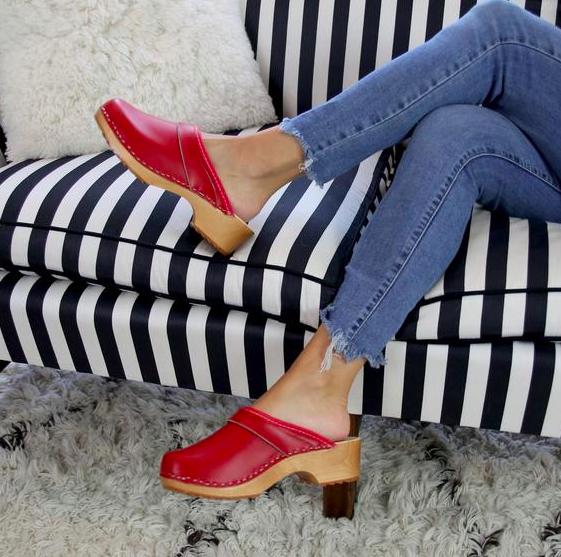 Model wears  distressed jeans and slip-on clogs with a bright red upper and a light wood heel while sitting on striped couch