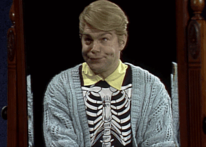 a gif of stuart smiley making faces at himself in a skeleton shirt with a cardigan over it