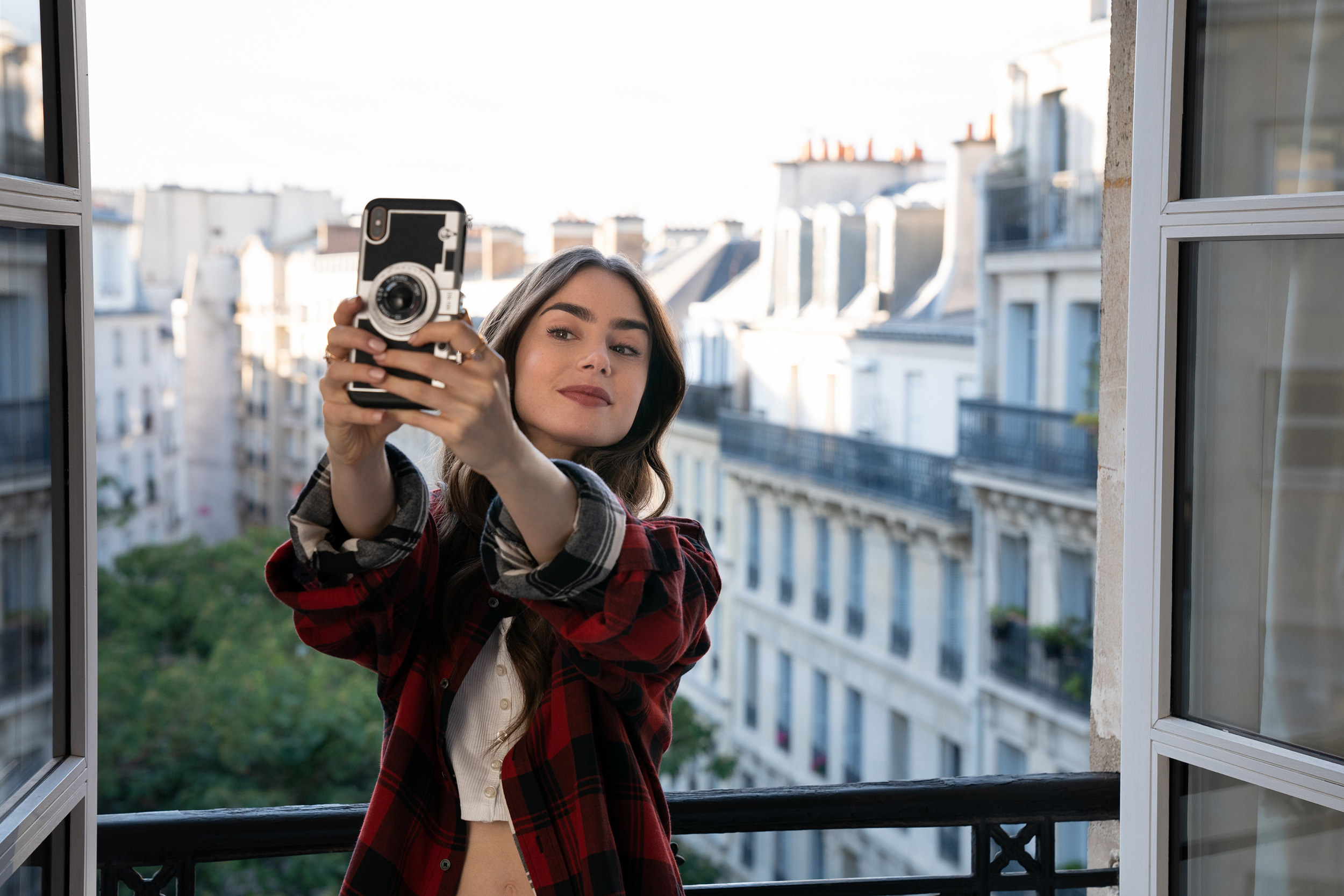 Cena de "Emily em Paris", Lilly Collins tirando uma selfie numa janela. 