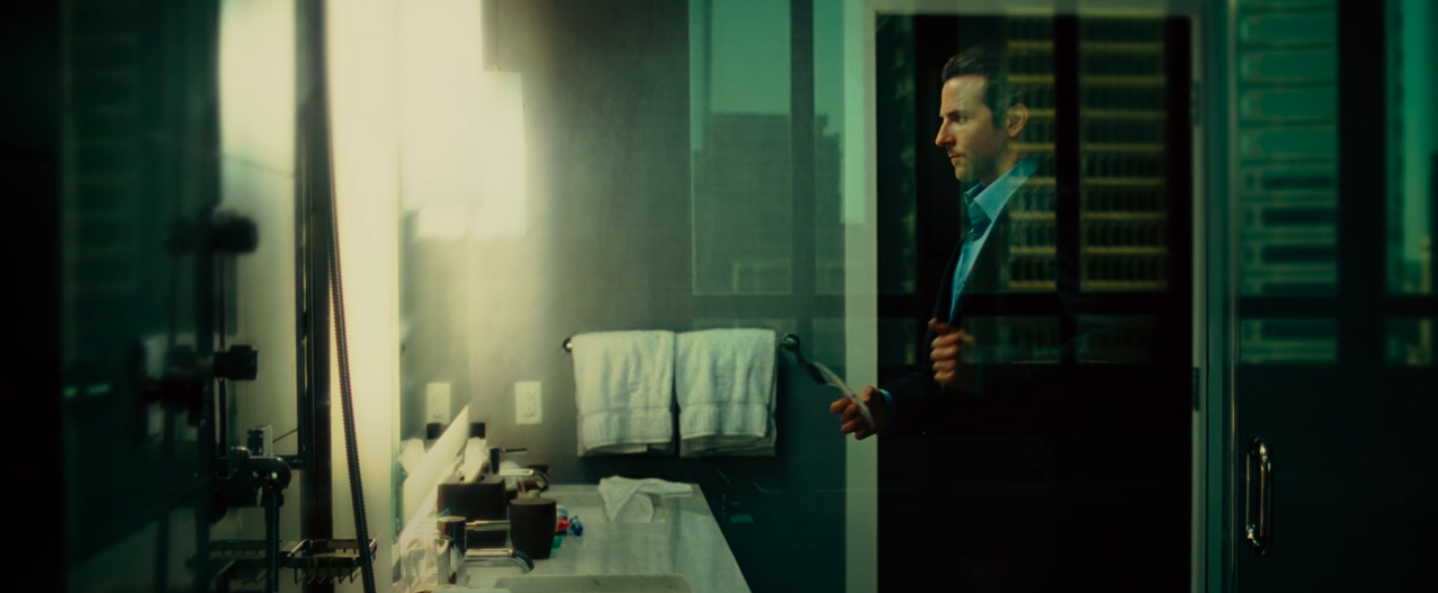 Bradley Cooper stands in the mirror wearing a suit and a shirt with an extremely high collar