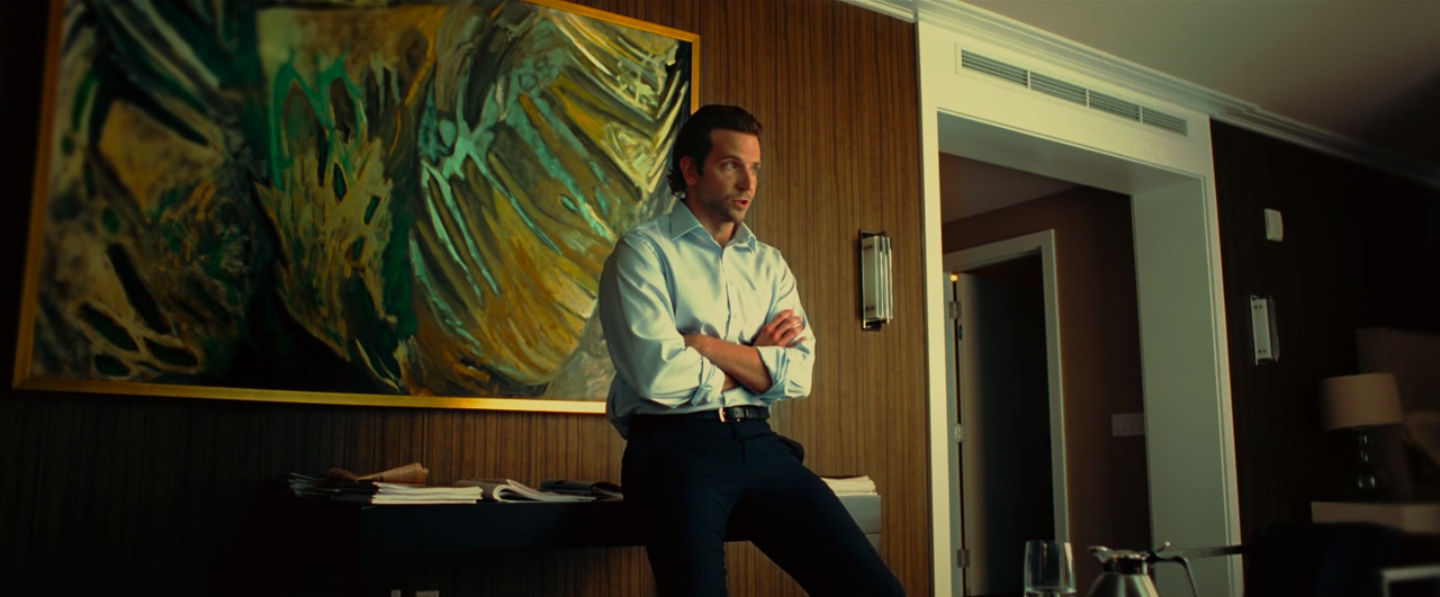 Bradley Cooper in a shiny button down and slacks, barely sitting on a desk