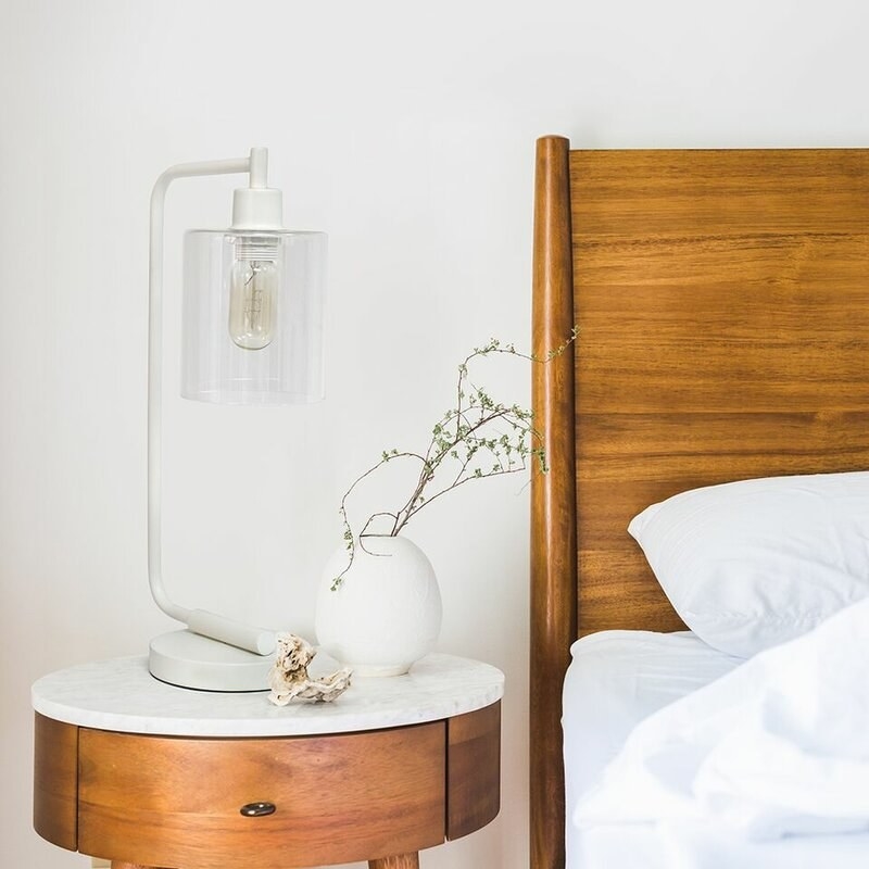 A bedroom side table with a lamp and bed with white sheets