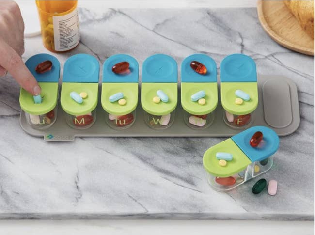 A set of seven transparent pill bottles with blue and green silicone lids dividing day and night propped on a gray magnetic holder 