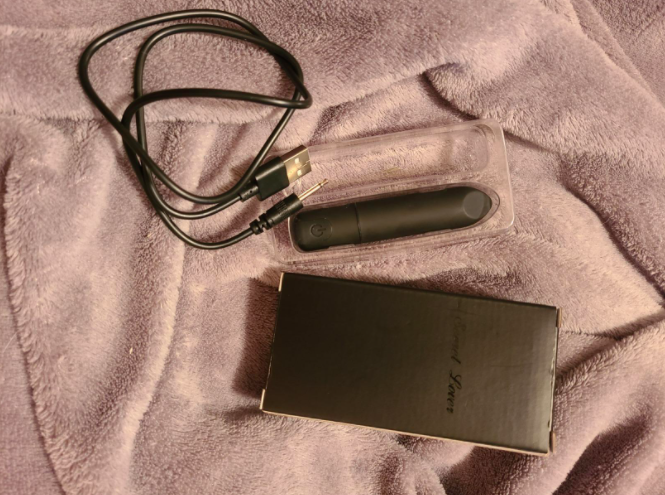 The Bullet Vibrator with Angled Tip and its charger and packaging on a customer&#x27;s bed