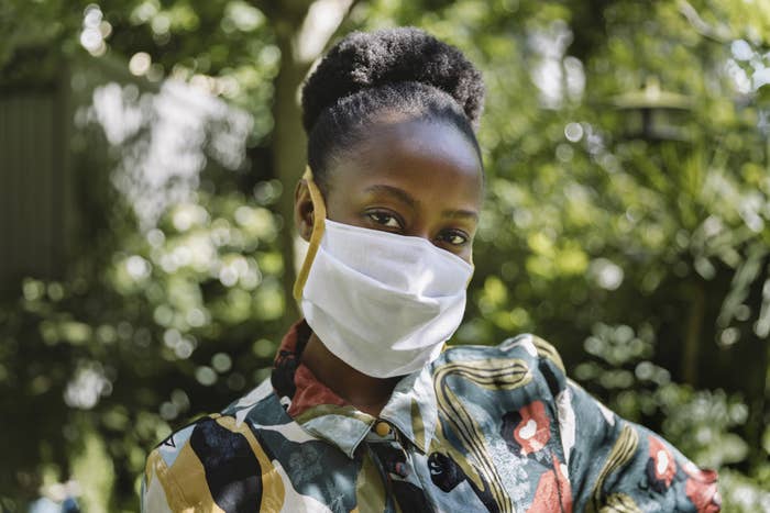 A person against a leafy backdrop, wearing a pleated mask and a printed blouse.