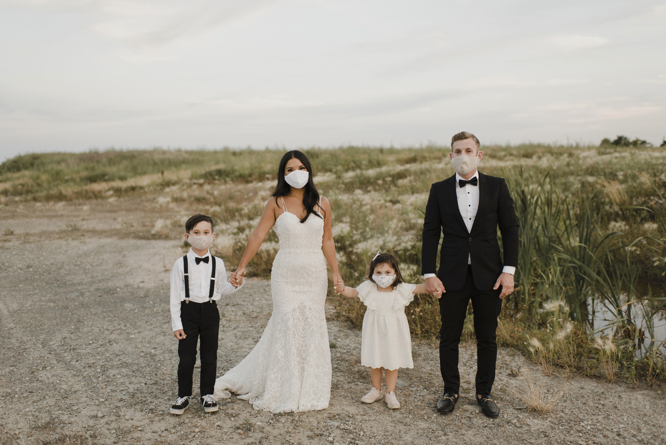 A bride and groom wearing face masks while holding the hands of two children, also wearing masks