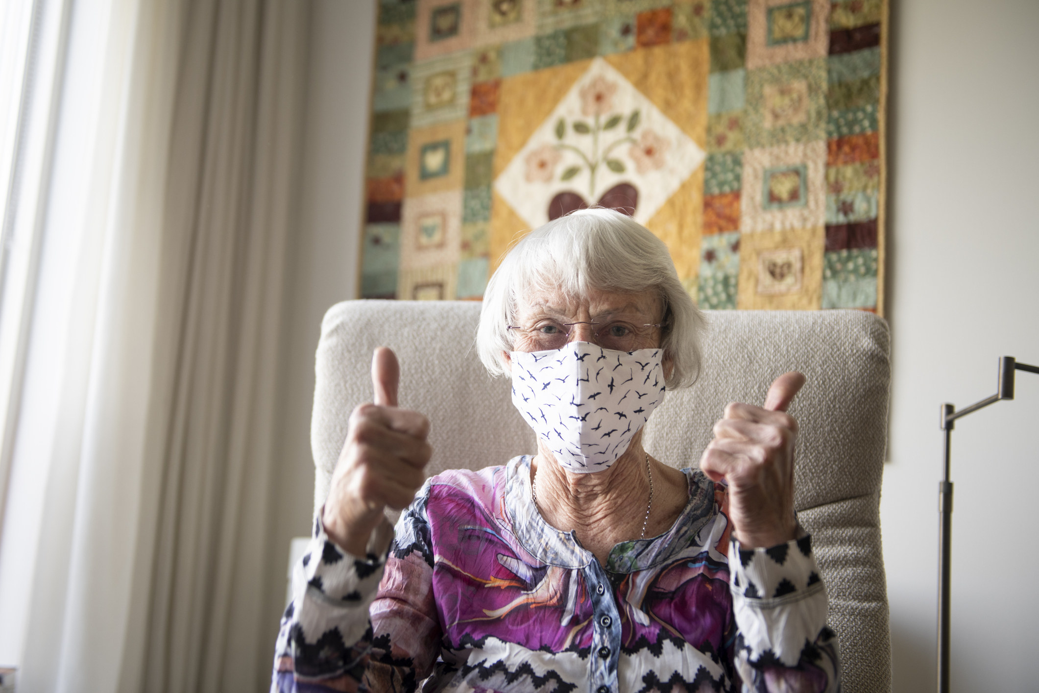 Une personne âgée assise dans un fauteuil et portant un masque qui lève les pouces.